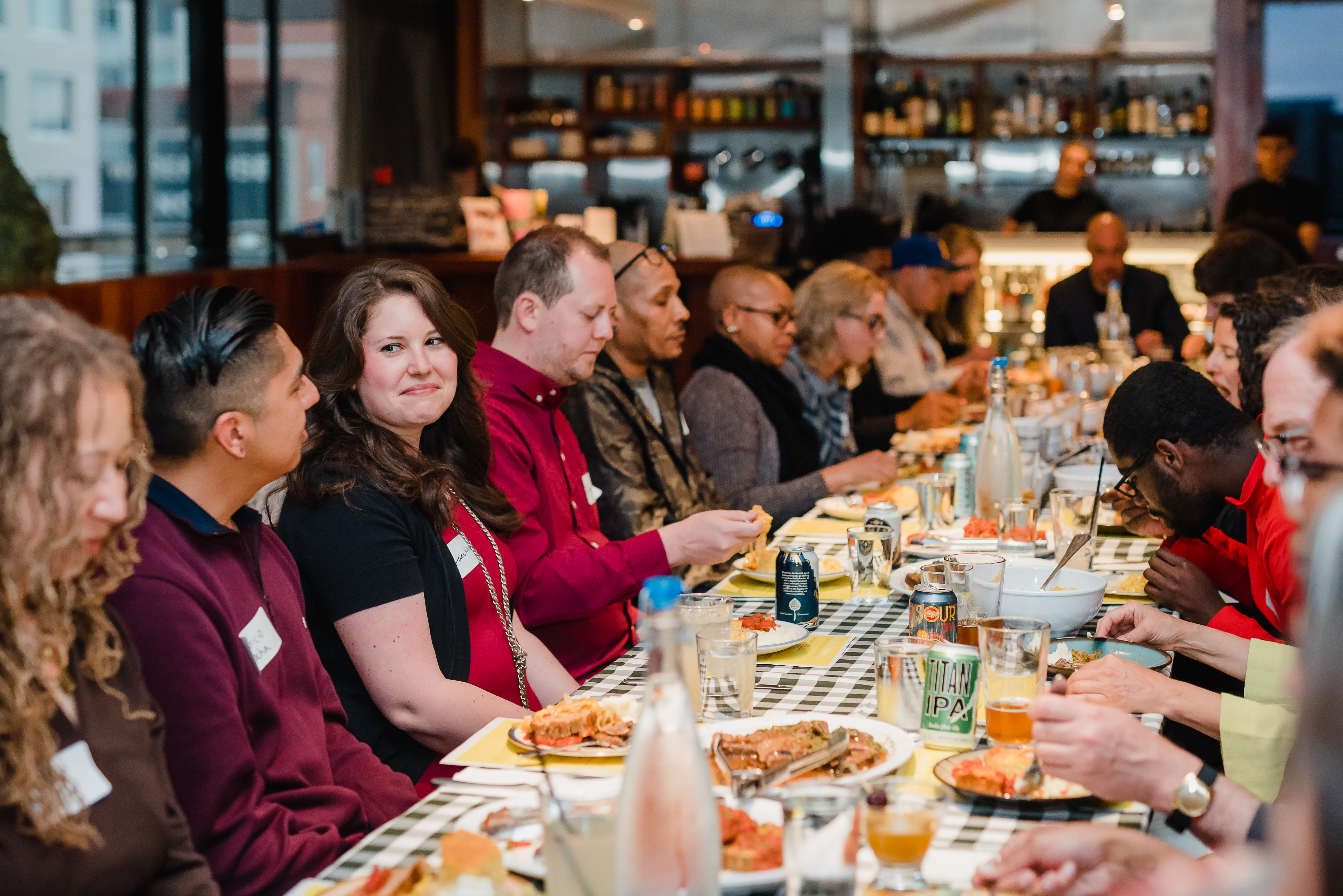 About two dozen people sit at a table eating food and engaging in conversation. The crowd is diverse in ethnicity and age. 