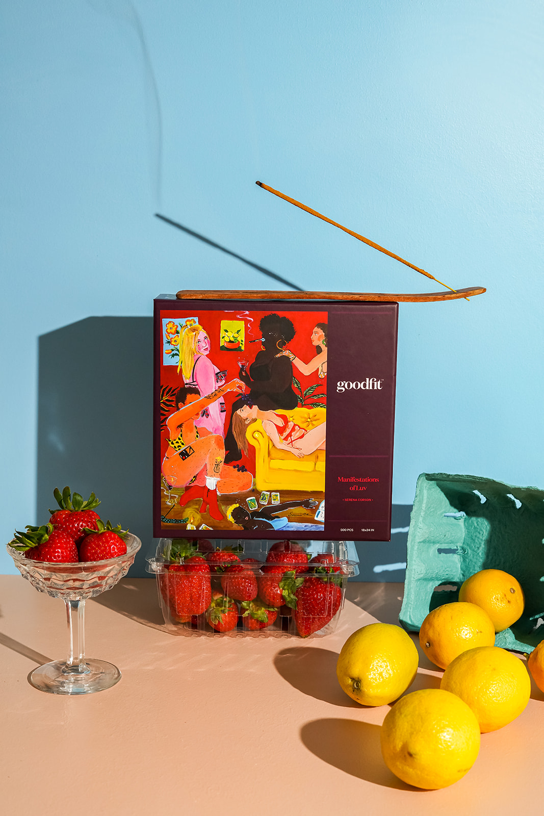  Puzzle box sitting on a clear container filled with strawberries. To one side of the puzzle box there is a coupe glass, also filled with strawberries. On the other side is a container tipped over with lemons coming out. There is an incense holder and incense burning on top of the puzzle. The image on the puzzle is a vibrant portrayal of six figures drinking, smoking, feeding one another, and laying on the floor. 