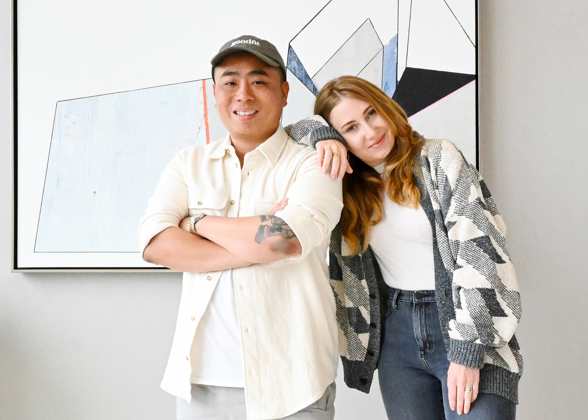 Bryan and Casey of Goodfit Puzzle standing next to one another in front of a geometric work on art. Bryan’s arms are crossed and he is sporting a button up and a baseball cap that says “goodfit.” Casey has her arm resting on Bryan’s shoulder and her head resting on her arm. She is sporting black jeans, a wide t-shirt, and a cardigan. 