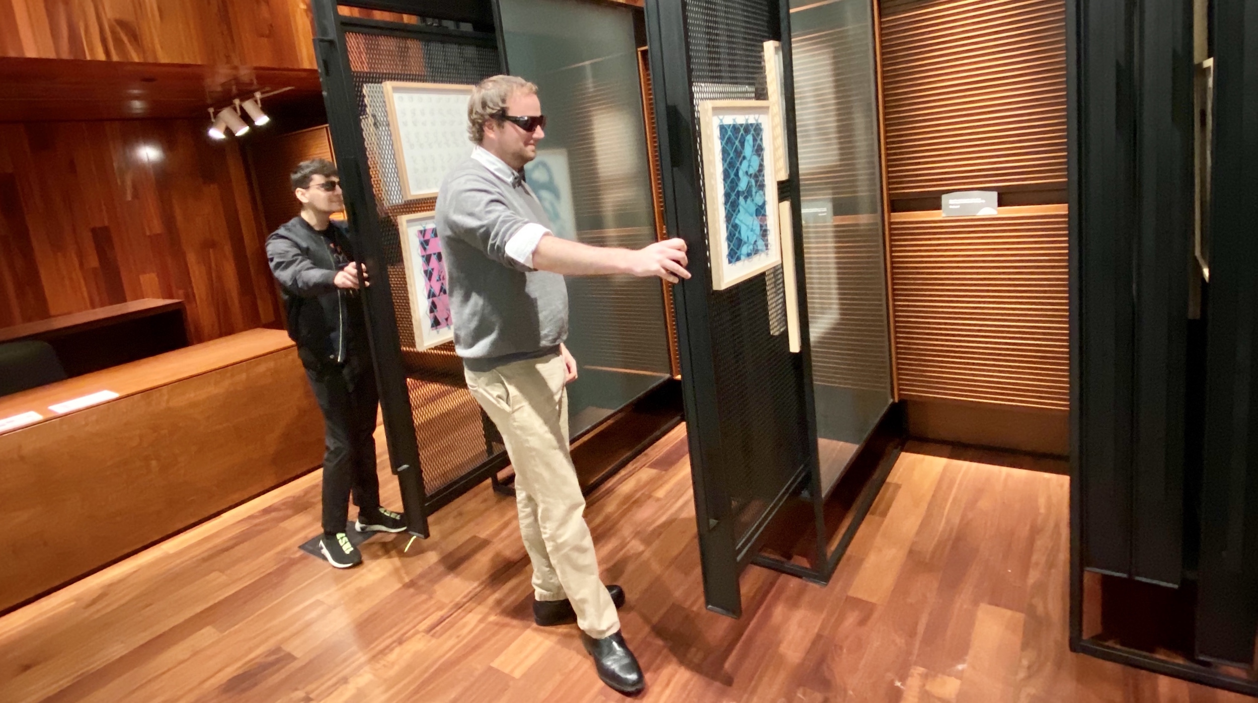 Two young men wearing color correcting glasses view artworks in a gallery and are smiling brightly. 