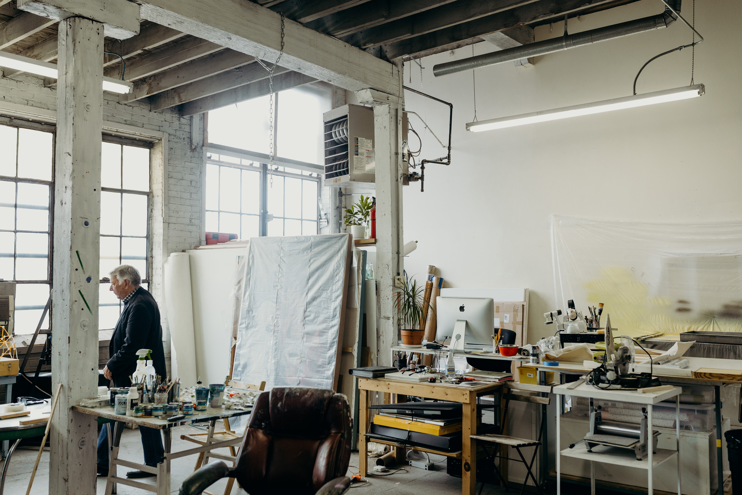 A large studio space. Clark is walking between tables that are filled with paints and brushes. 