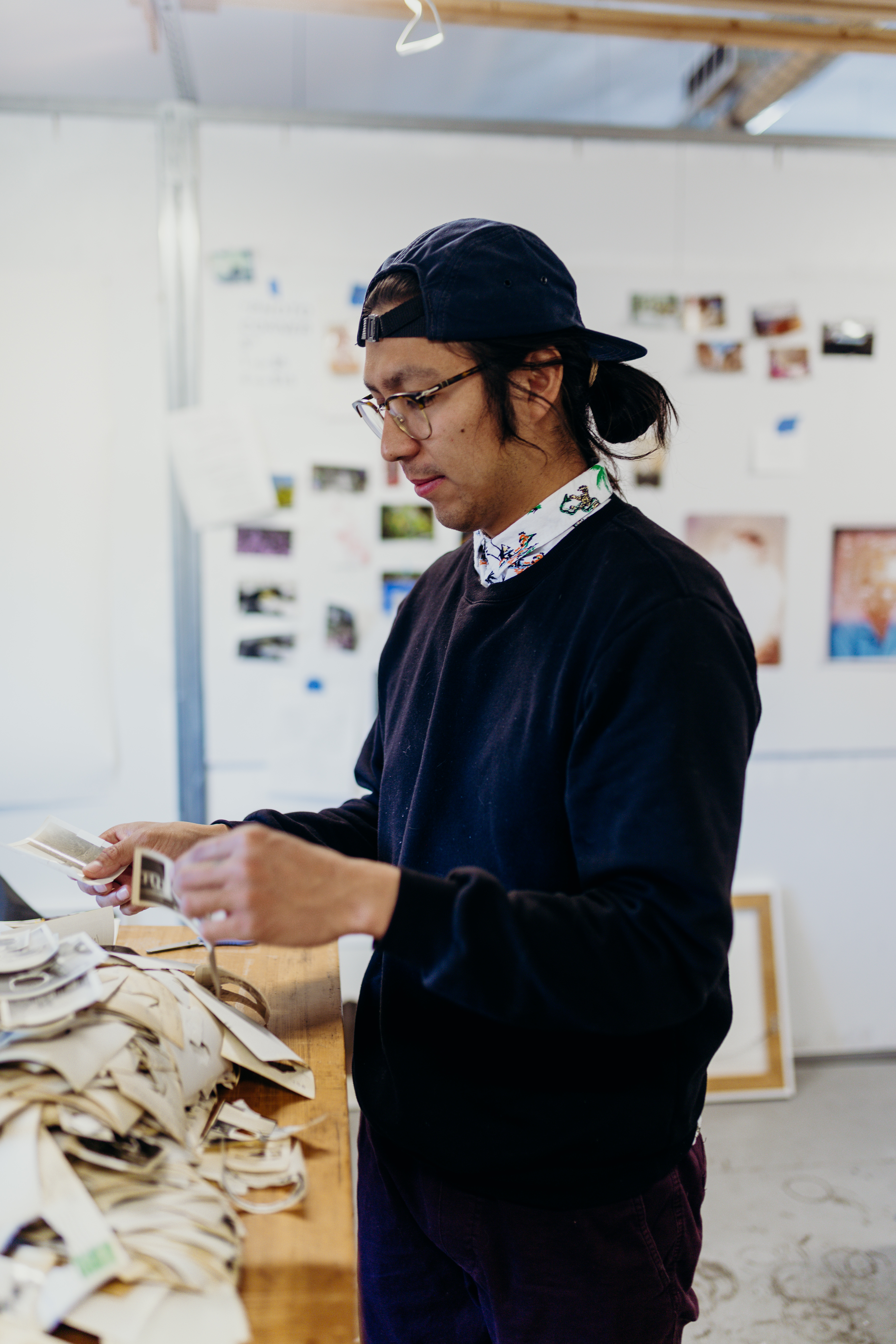 Image of George P. Perez in his studio looking at photographs. He is wearing glasses, a blue sweater, jeans, and a blue baseball cap. 