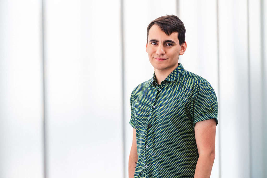 headshot of Dylan in one of the MCA Denver white galleries. He is wearing a short sleeve green button up, with diamond shapes on it. His ams are to his side, and he’s looking directly at the camera with a soft but sly look on his face. 