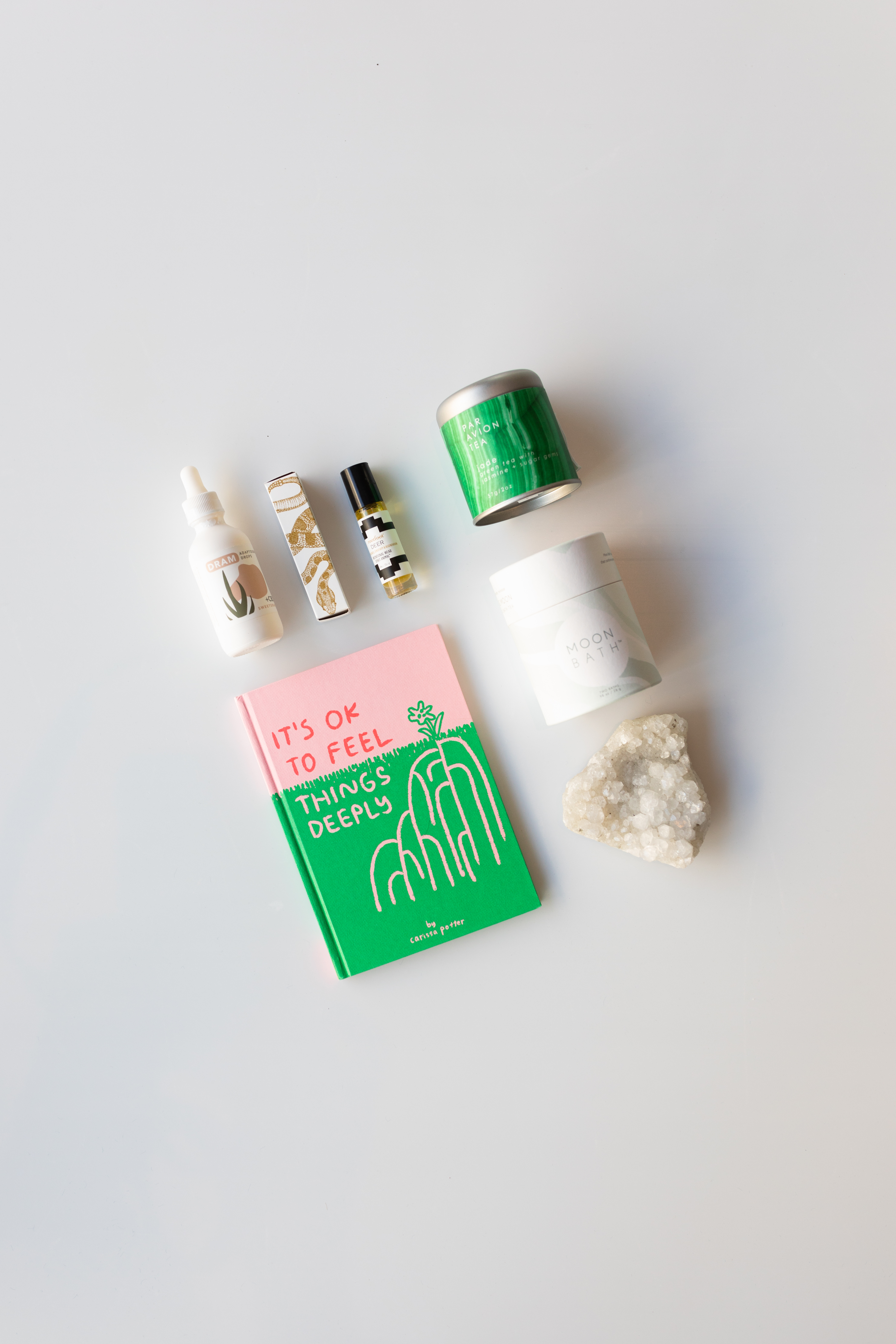 A hotpink and bright green book reads It’s ok to feel things deeply; A white crystal rock; a vial of cbd drops from Dram. Green tea, jasmine, and sugar jems mix from Par Avion Tea; a white container reading moon bath; a scent roller from rootfoot. 