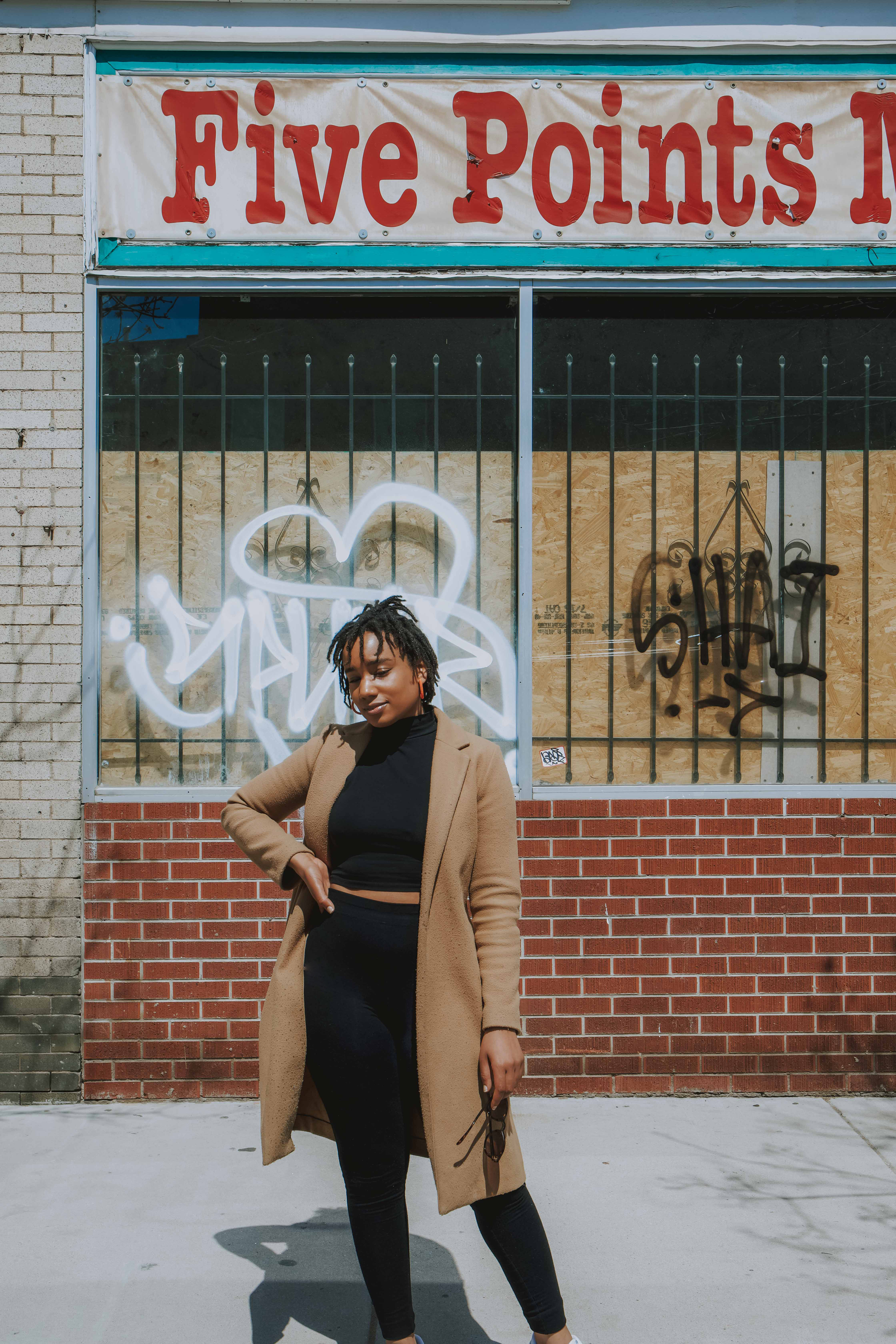 Artist, Narkita Gold, Standing on a street in front of a store with the words "Five Points" written in red on the marquee of the store. She is in a camel colored coat and black mid drift shirt and black pants. Her hand is on her hip and her head is at a profile with her chin down and eyes looking towards the ground. There is a soft smile on her face. 