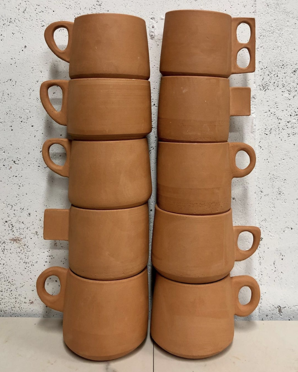 Two stacks of terra-cotta colored mugs waiting to be glazed. Each stack includes five mugs, and each mug has a different style handles on them. The mugs are sitting in front of a speckled black and white backdrop. 