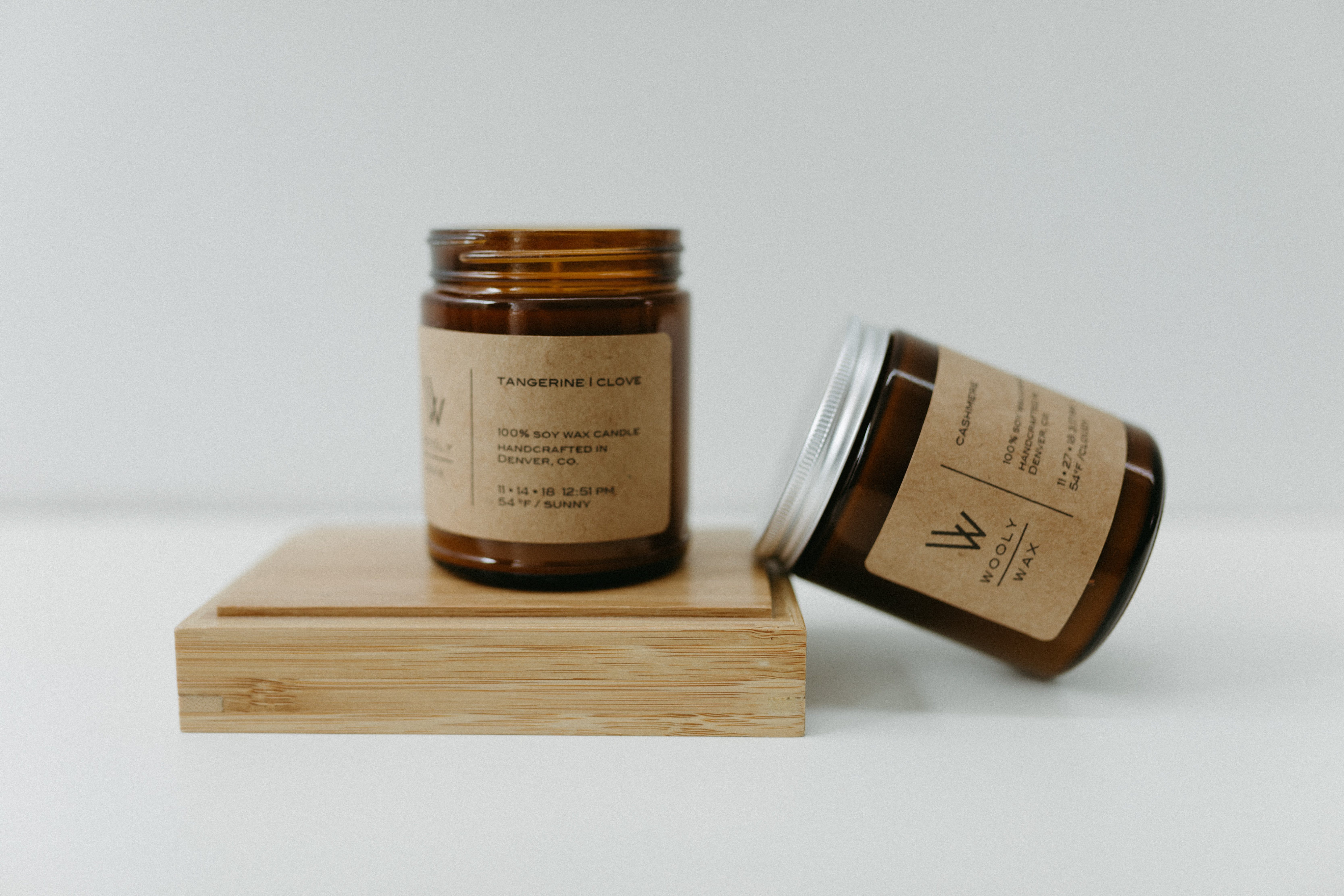 A tangerine and clove scented candle from Wooly Wax Candles is on a wooden platform against a white background. A second candle is tilted against the wooden platform.