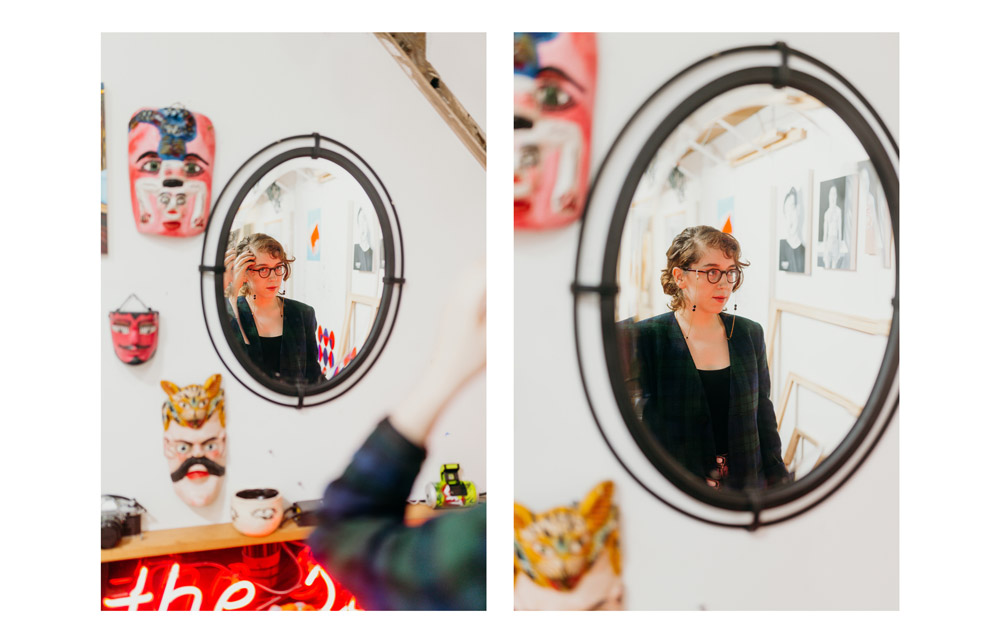 Sierra is seen through the reflection of an oval mirror hanging on a white wall. She is adjusting her hair behind her ear. 