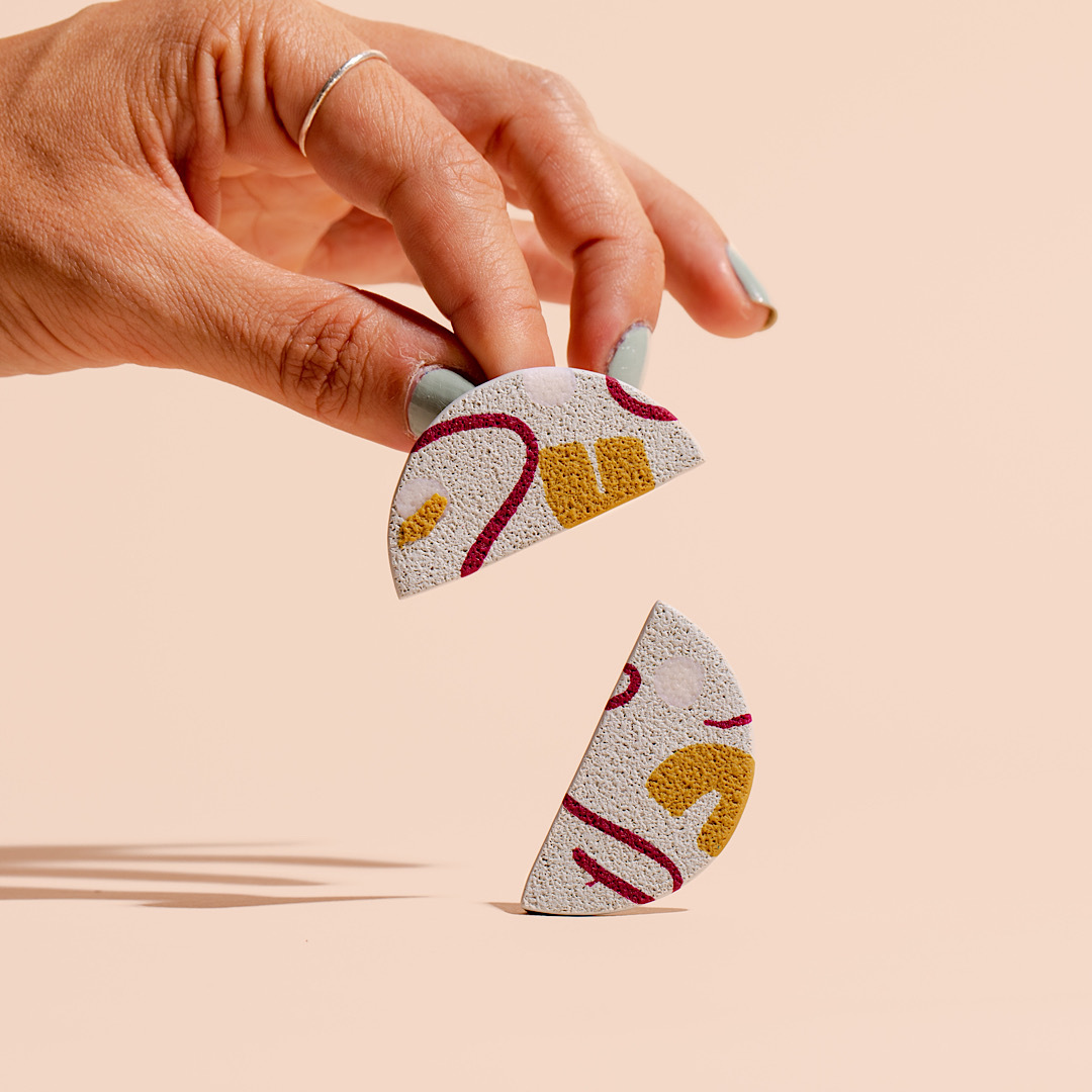 Two earrings, designed in the shape of a half circle, with red, yellow, and white geometric shapes on them. One earring is placed on the light peach colored background, and the other is being held by a hand. 