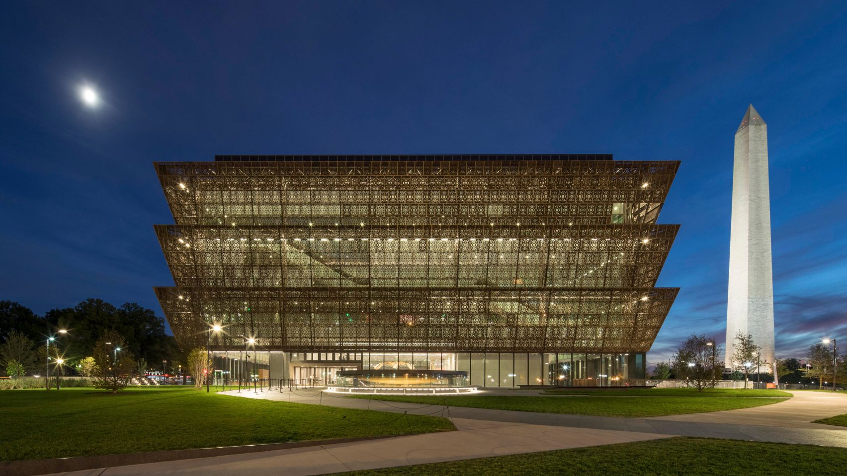 National Museum of African American History and Culture at night. The building glows from within. The Washington Monument stands in the background. 