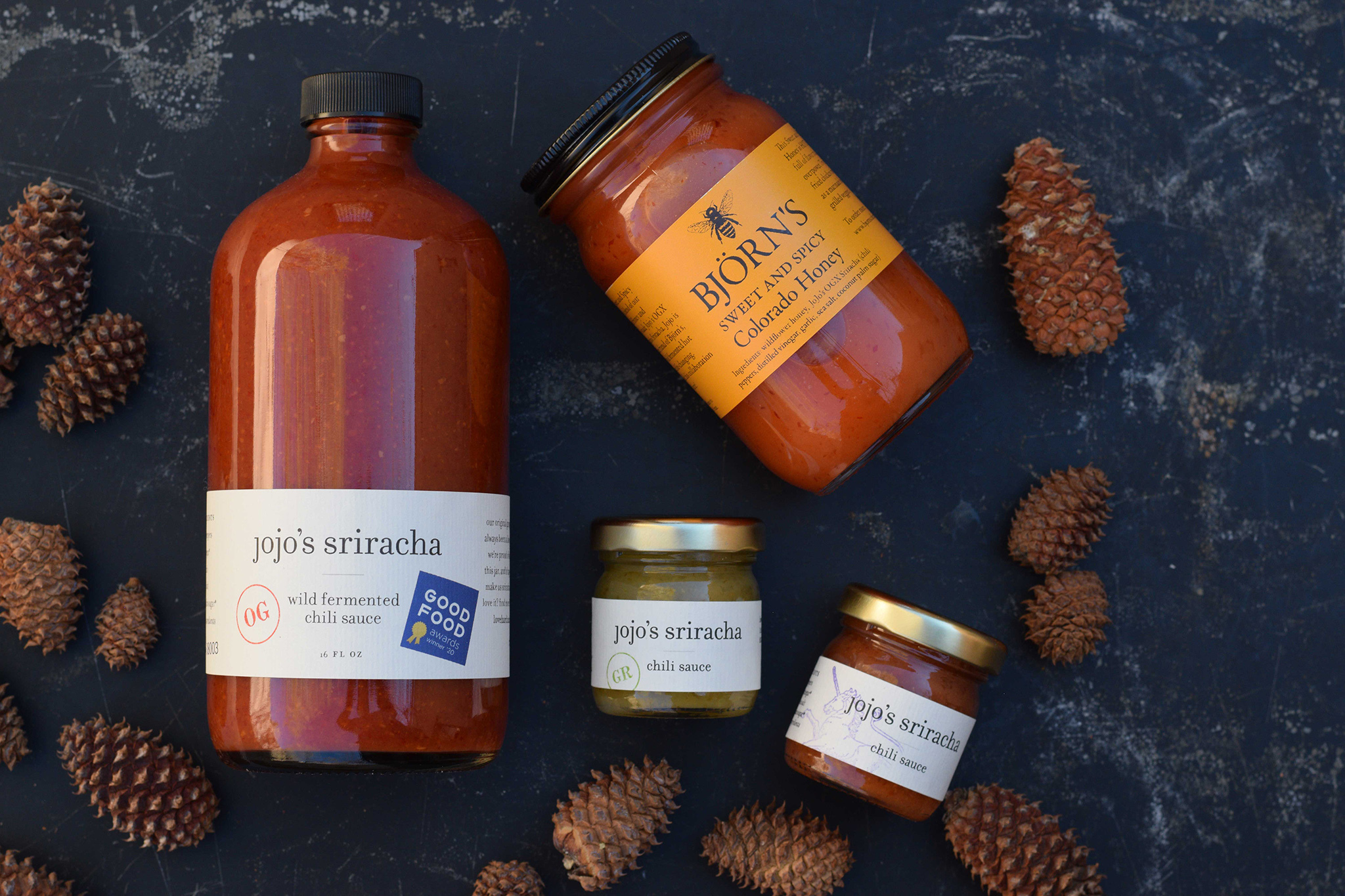 Four jars: three with sriracha inside, the fourth with honey inside (that looks like sriracha in color). The jars are photographed from a bird’s eye view and surrounded by pine cones.