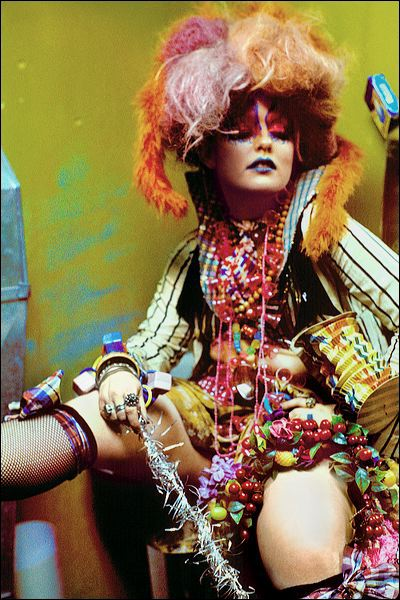 An image of a woman wearing colorful jewelry and clothing against a yellow background. 