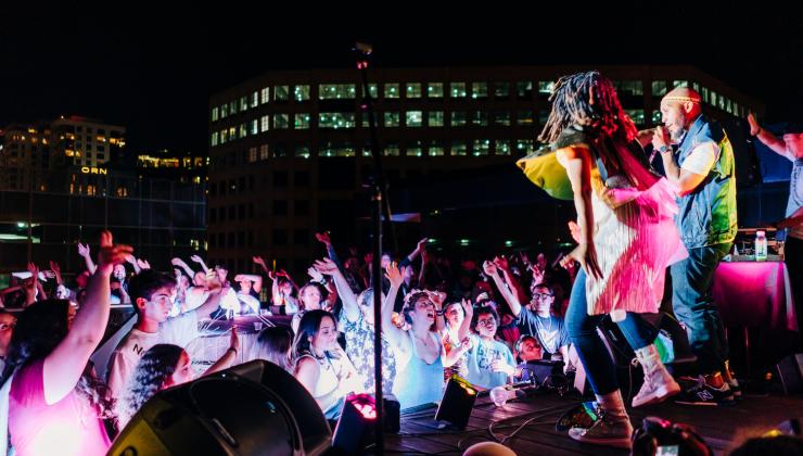 Two men are on a stage performing for a group of people at night at an outdoor space. The audience is washed in a pink light.