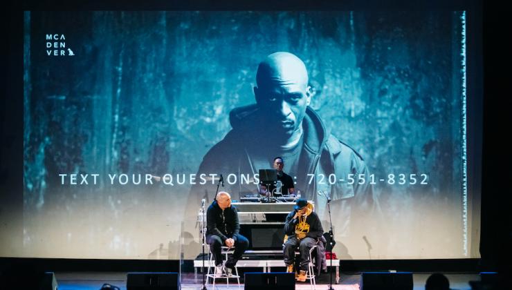 Rakim is sitting on a concert stage next to another man. Behind them is a projection of Rakim looking into the camera with the MCA Denver logo in the upper left corner. 